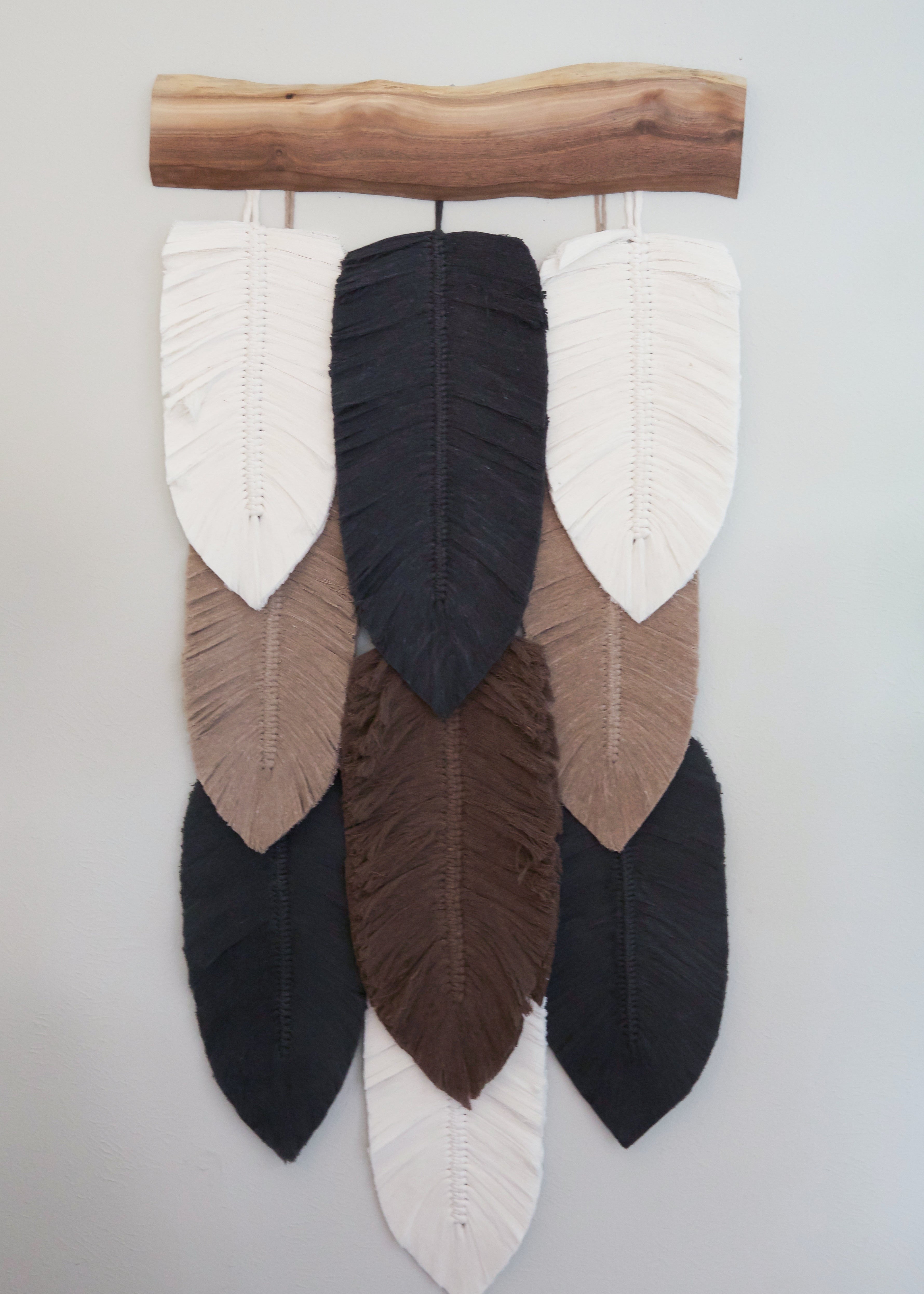 A wall hanging decor featuring a series of handcrafted macramé leaves in shades of beige, brown, and black, strung together on a wooden plank.